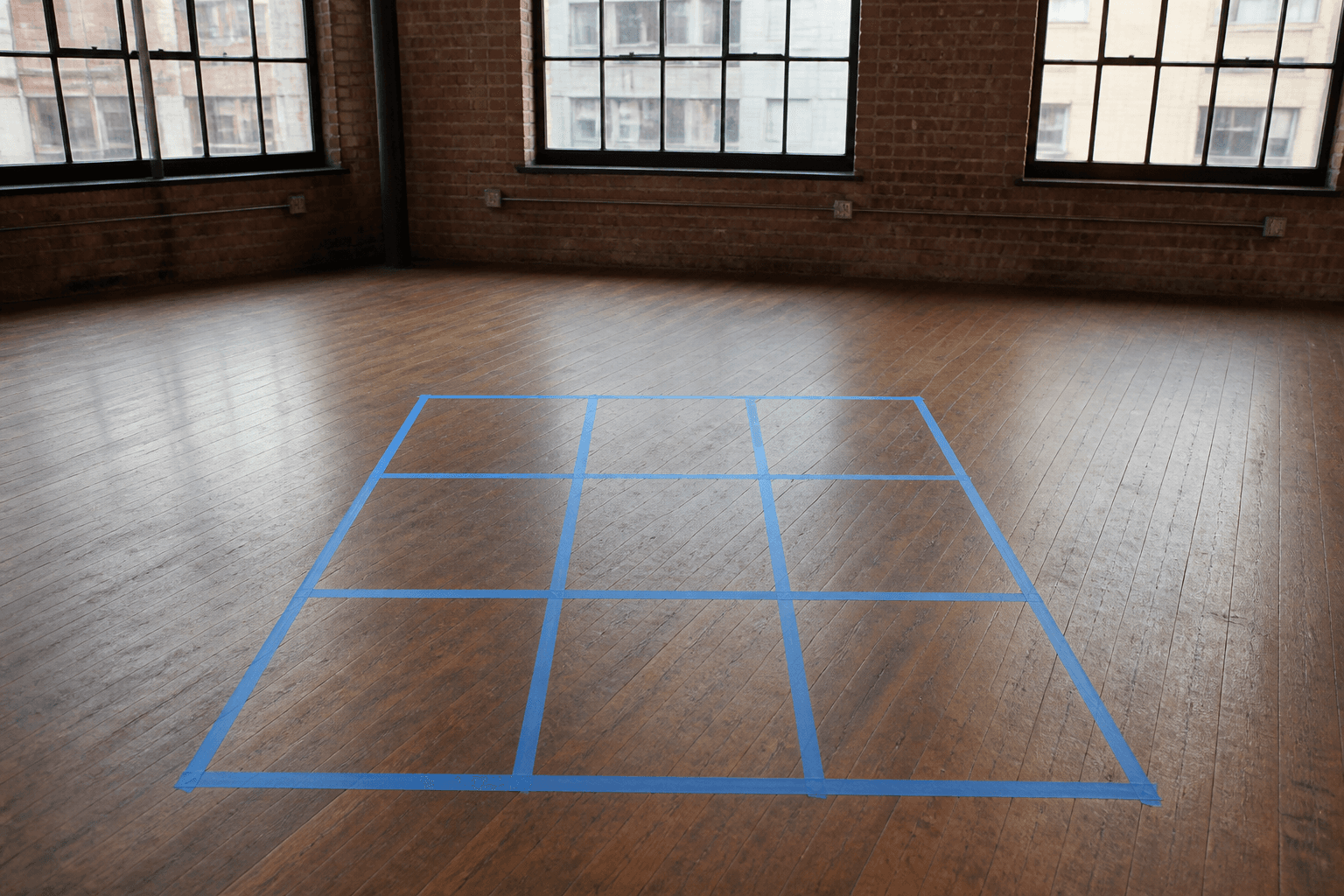 Blue painter tape in a 3x3 on a loft wood floor, tall windows in the distance