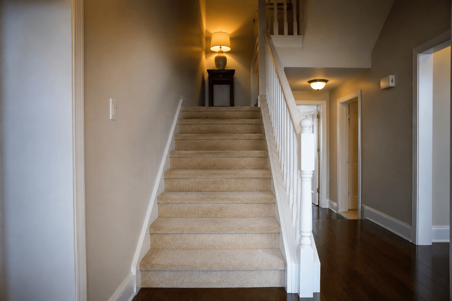 Townhouse stair from the bottom landing, beige carpet, white banister, lamp on a small table
