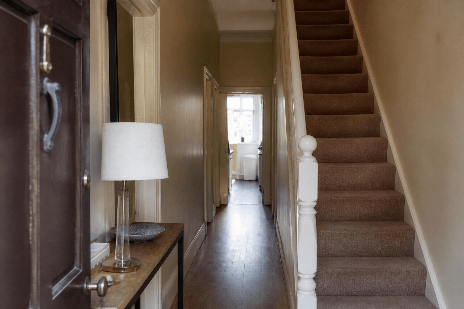 Long beige townhouse hall from the open front door, stair on the right, lamp on a slim console, feng shui bagua for townhouse