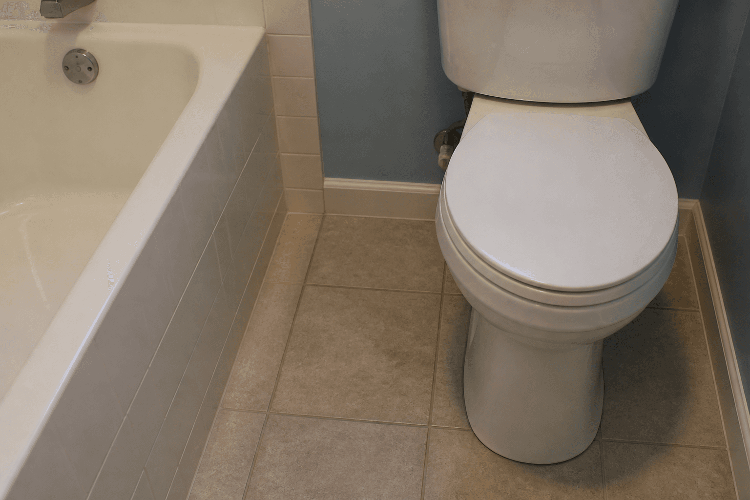 Closed white toilet lid beside a soaking tub, short tiled aisle