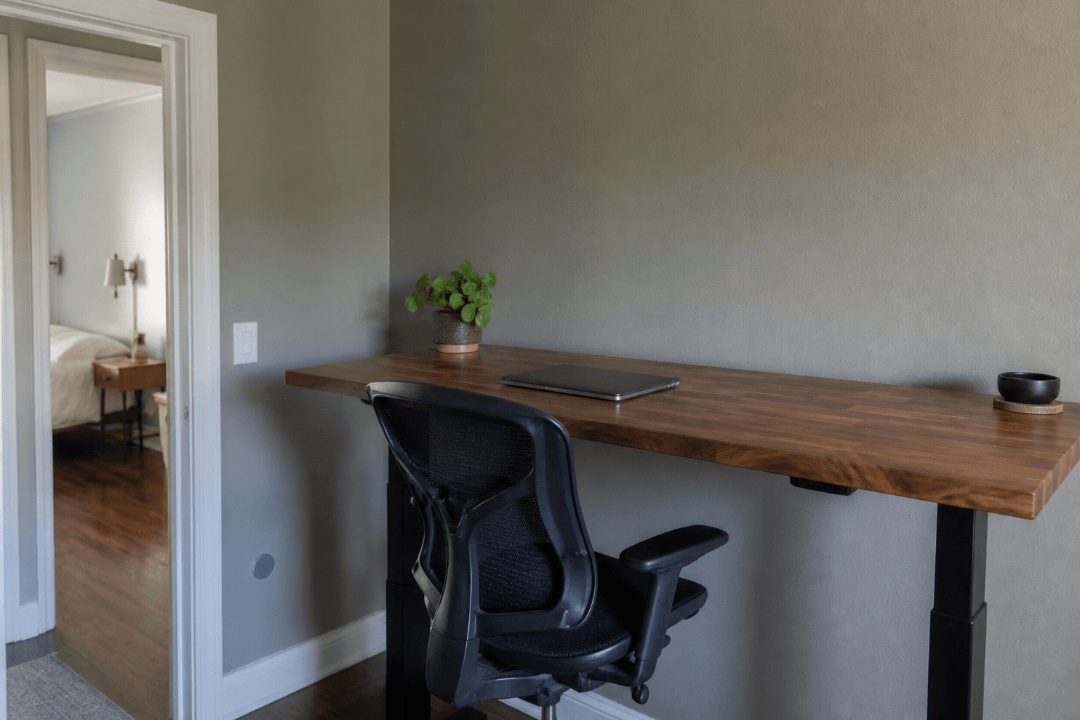 Closed laptop on a Feng Shui standing desk, chair tucked, earth-tone rental, doorway visible from the standing spot