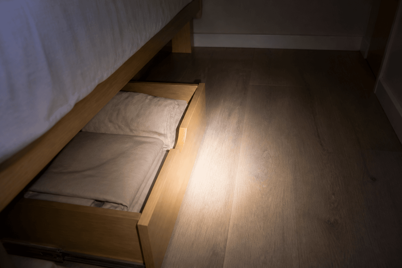 Open under-bed drawer with folded beige linens and a flashlight on the box
