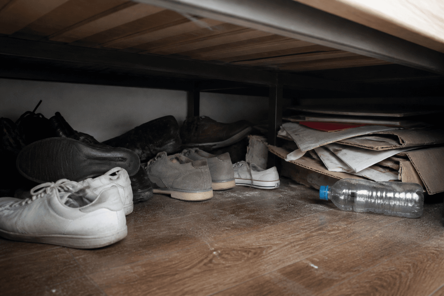 Shoes, a water bottle, and a cardboard pile stuffed under a platform bed