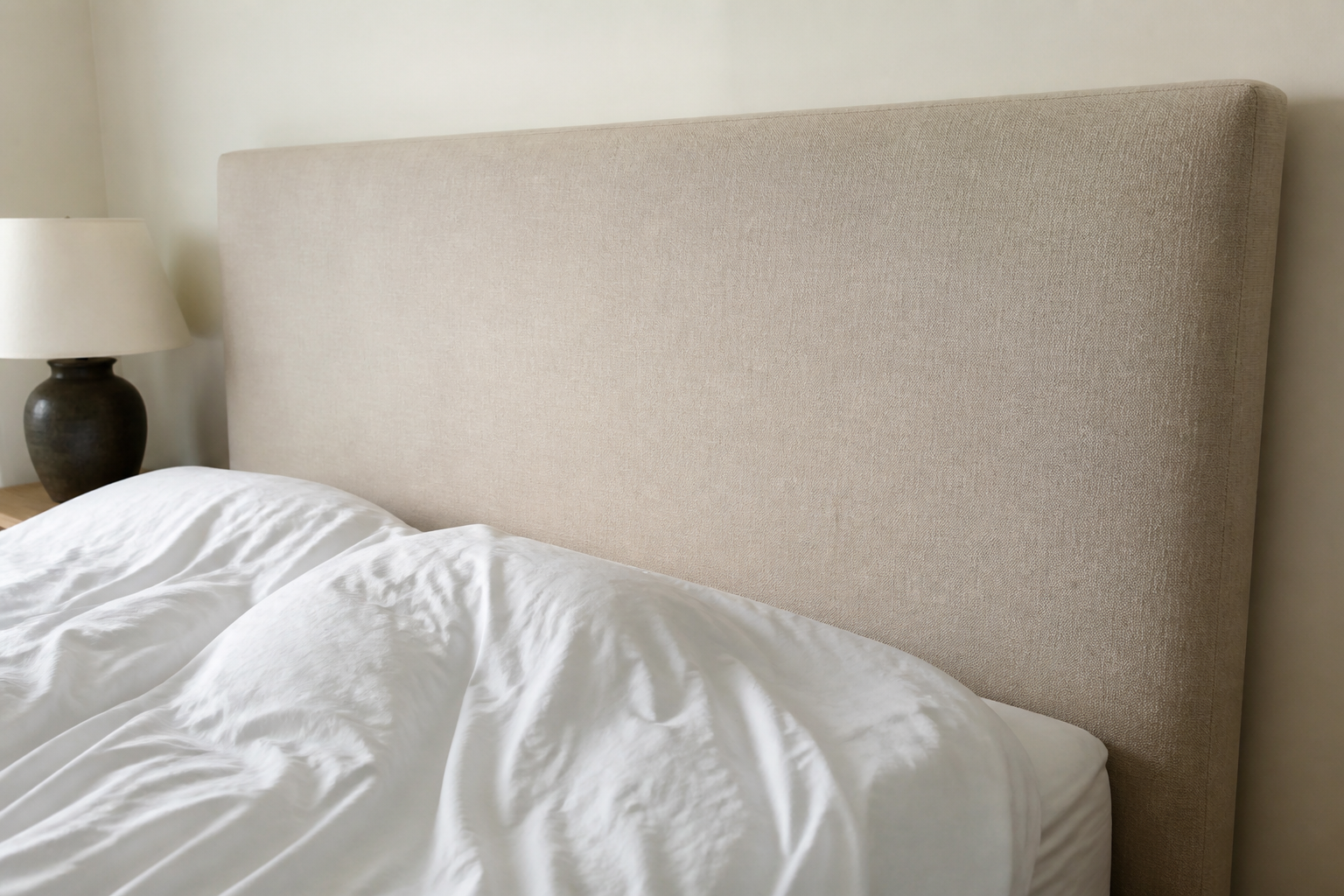 Tall oatmeal upholstered fabric headboard on a rumpled bed against a cream wall
