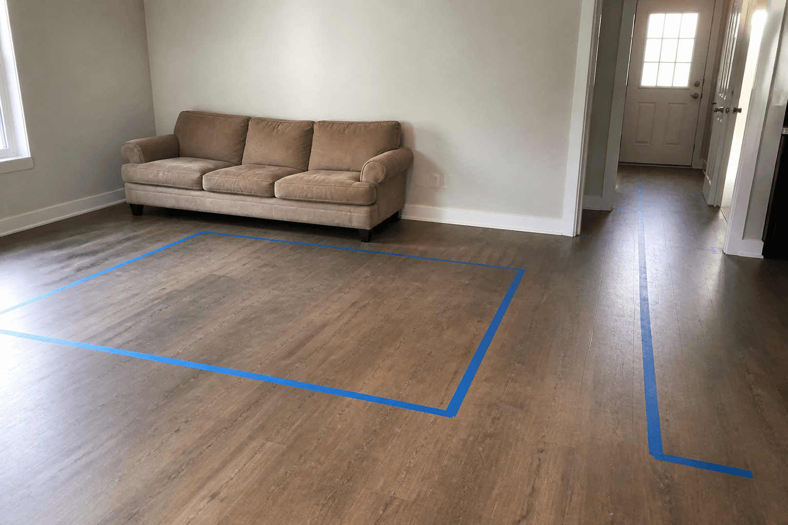 Blue painter tape on a wood floor marking a sofa footprint and a walk to a door