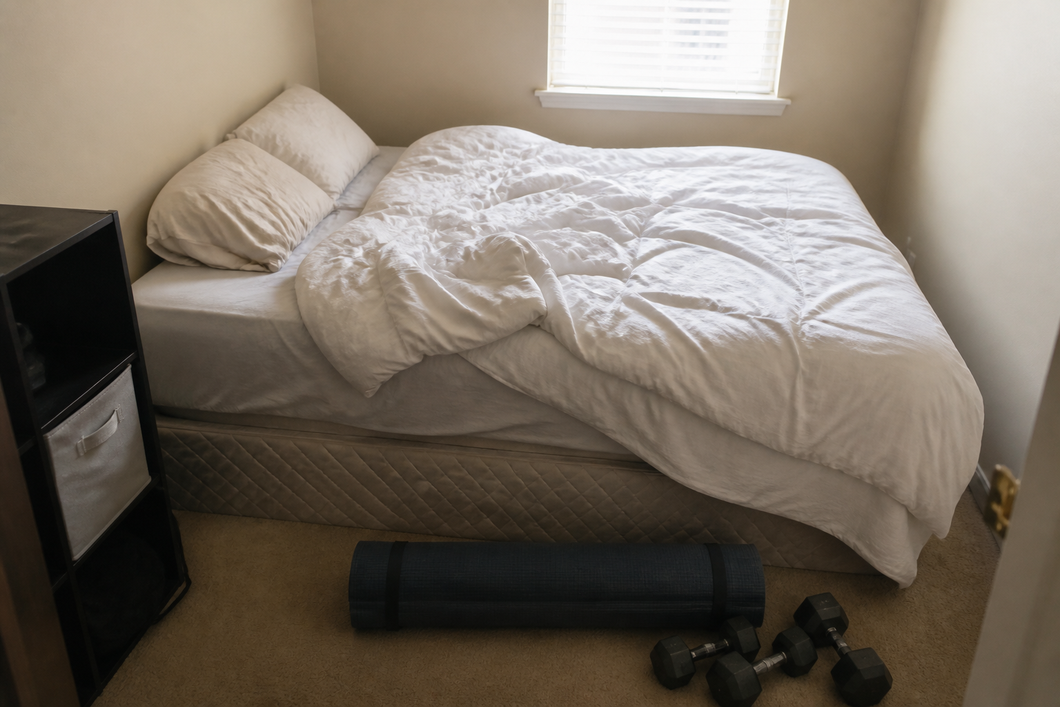 Yoga mat and dumbbells stored at the foot of a rumpled bed in a small beige bedroom