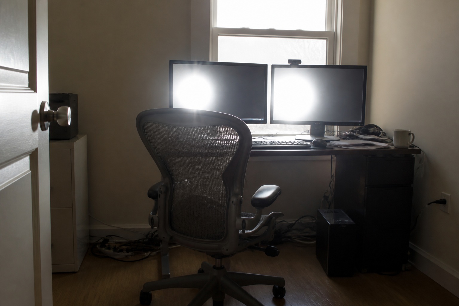 Two monitors catching window glare, chair with its back to the office door