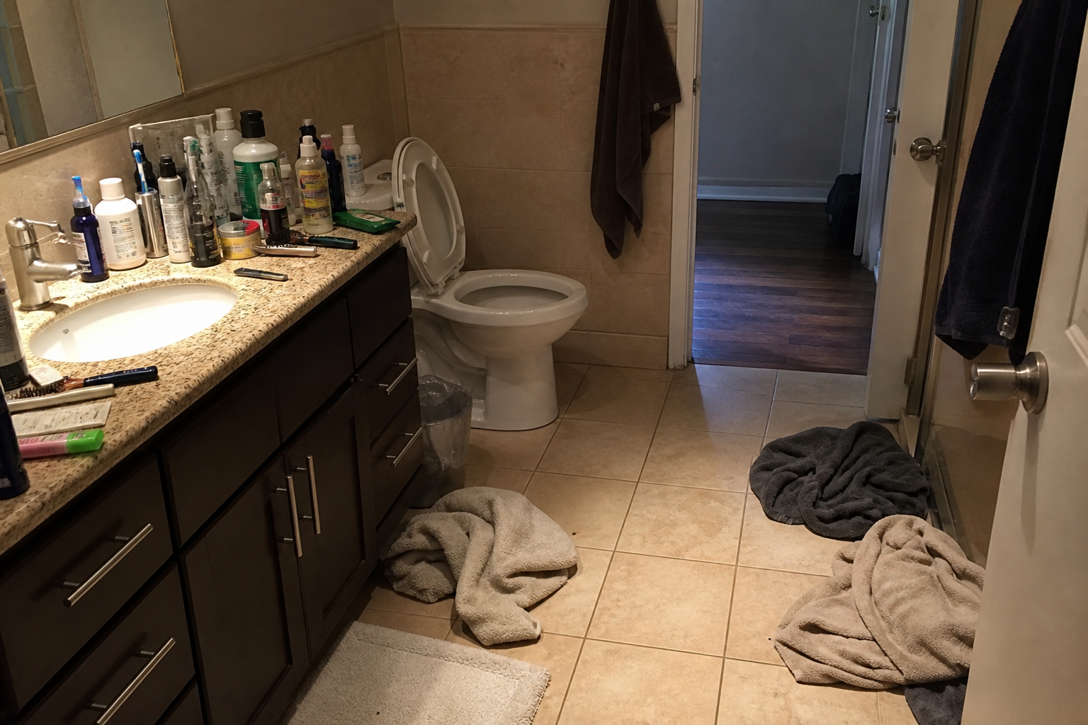 Crowded sink, open lid, towels on the floor, bathroom door open to a hall